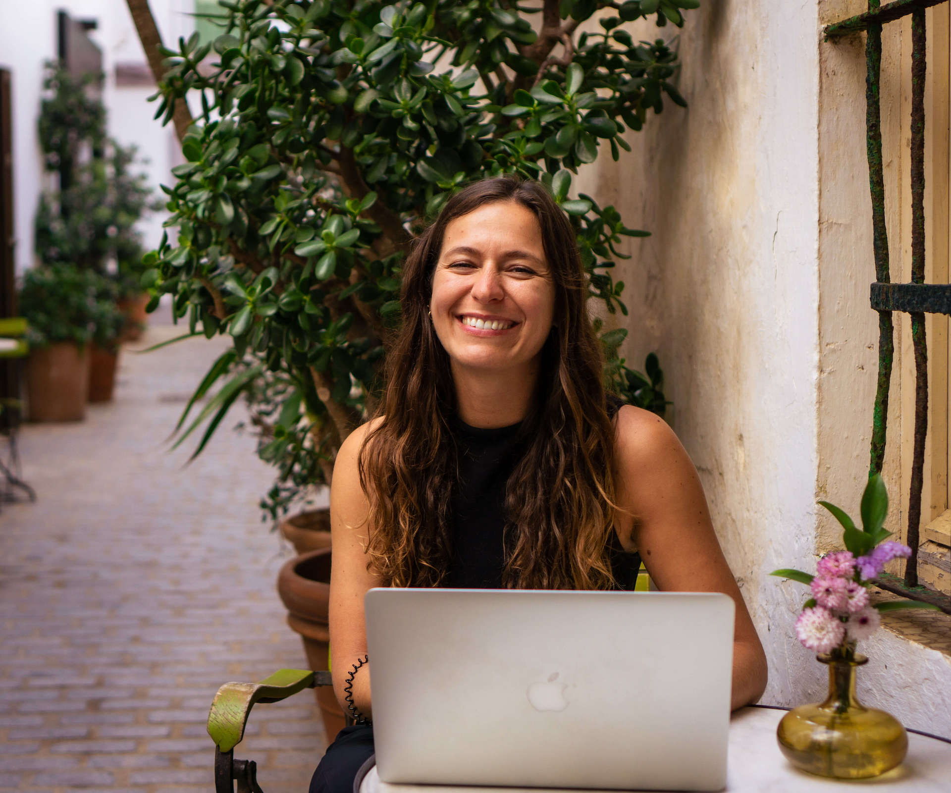 Frau sitzt im freien an einem Tisch vor ihrem MacBook und lacht