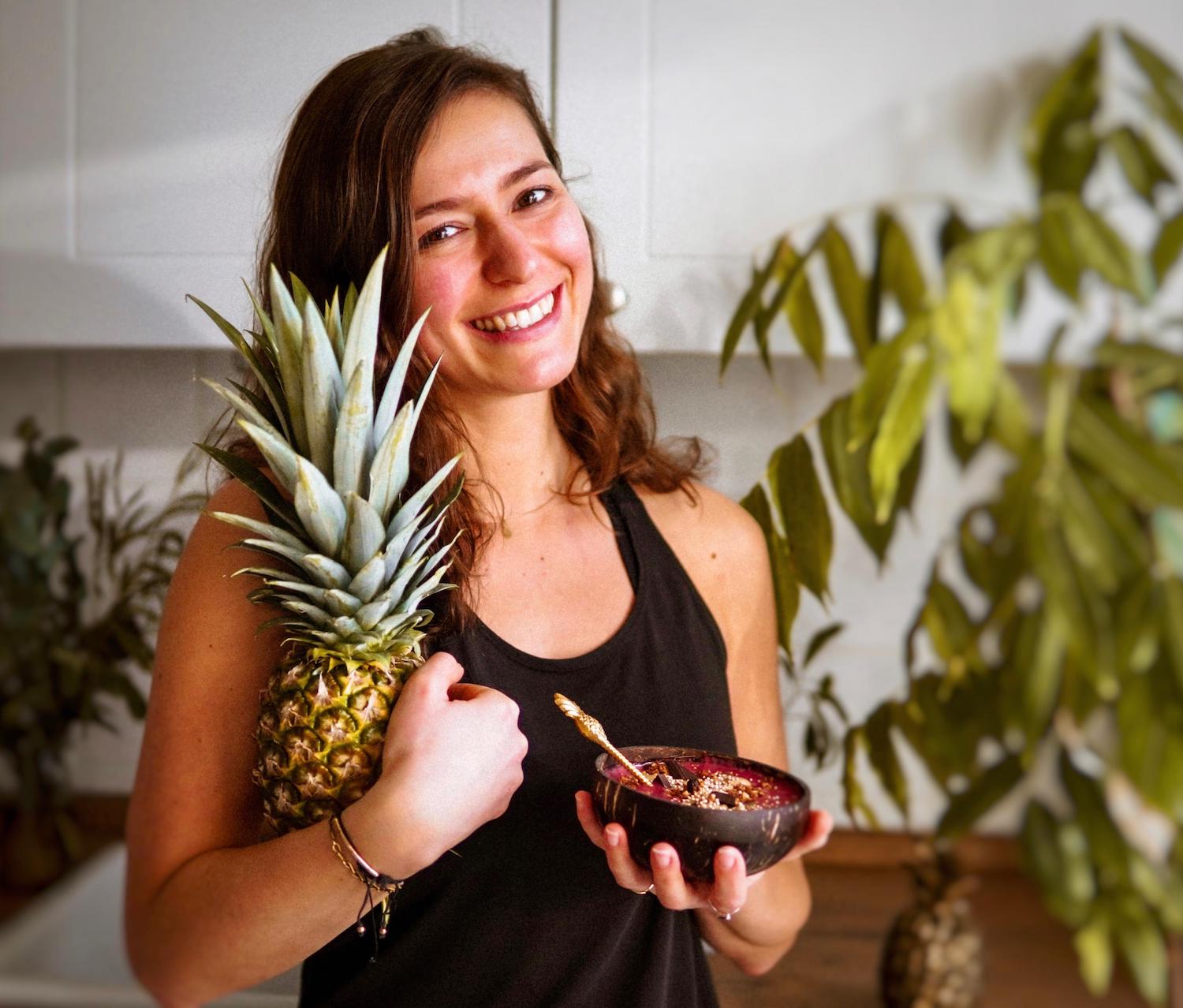 Frau hält links eine Ananas und recht ein Smoothie Bowl in den Händen und lächelt in die Kamera
